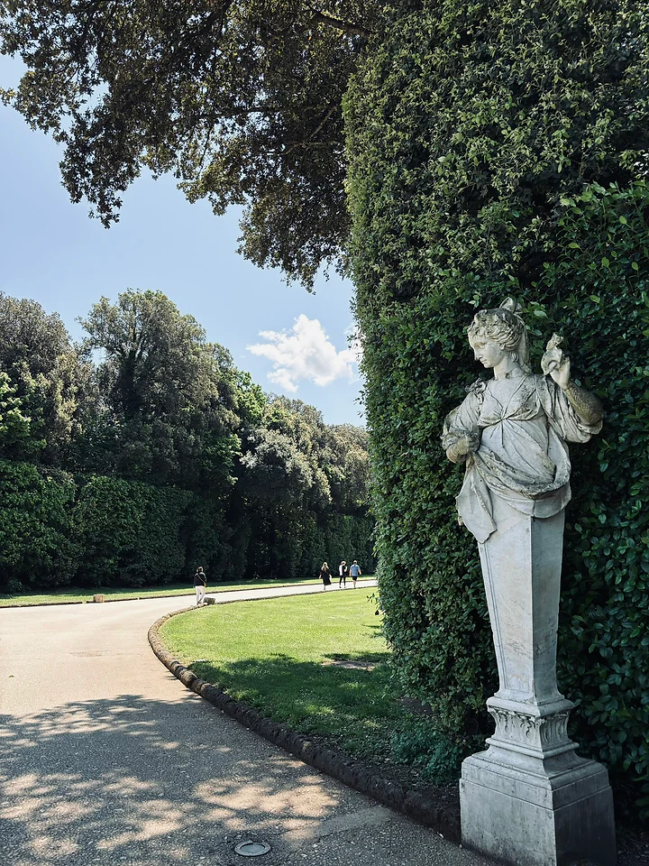 A weathered stone statue beside a clipped hedge in a formal garden.