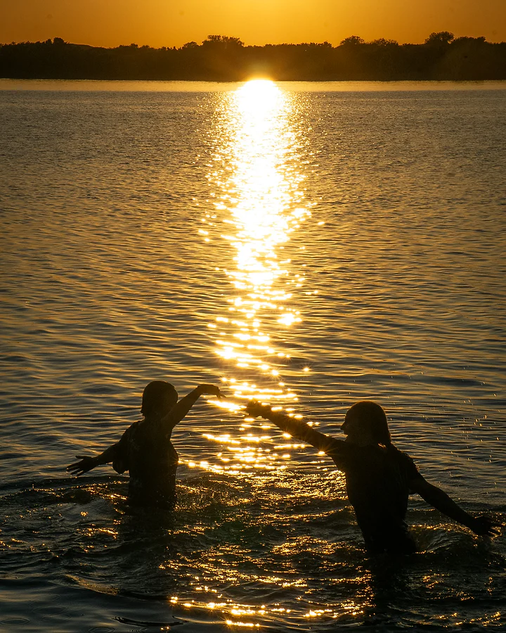 Two figures silhouetted in a lake, arms out, on a golden path of low sunlight.