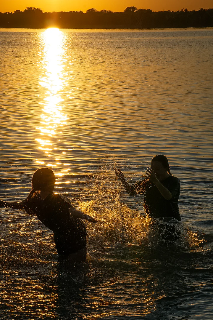 Two silhouetted figures splashing each other in bright golden lake water.