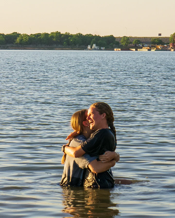 Two people hold each other, soaked, standing waist-deep in a calm lake.