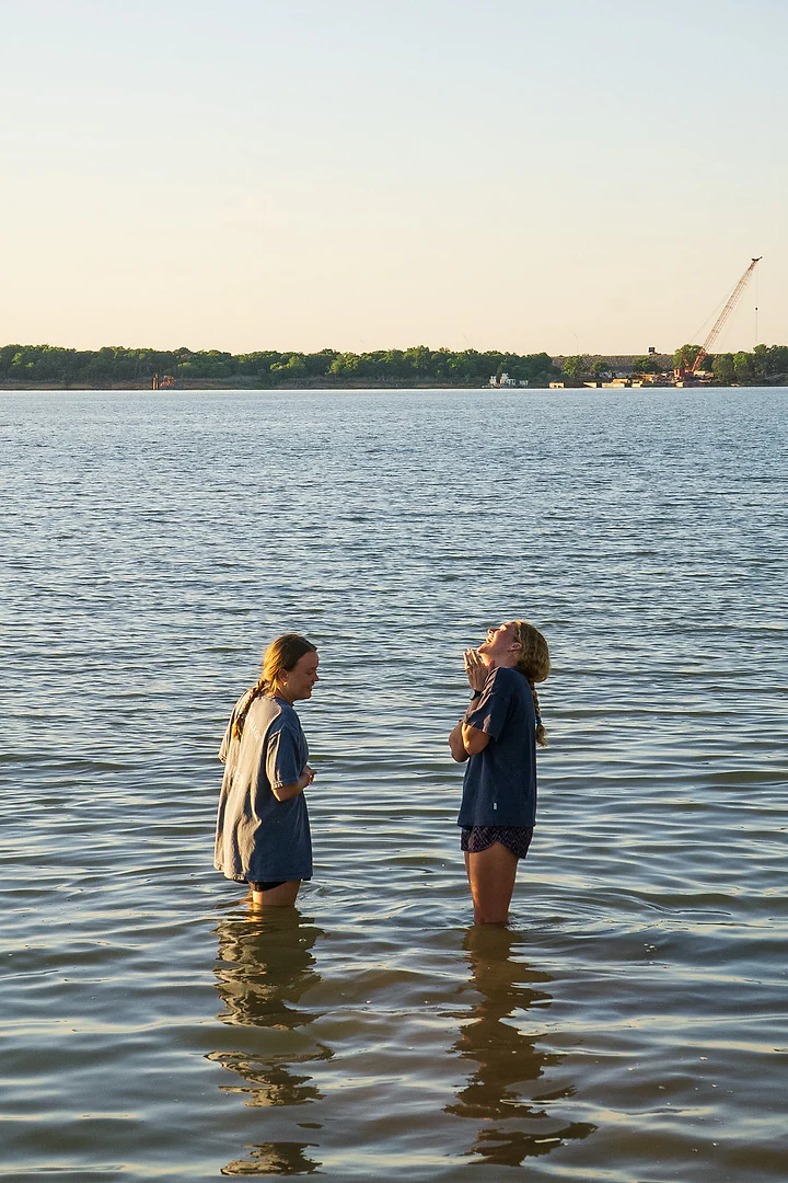 Two people standing knee-deep in still water, talking, under a pale evening sky.